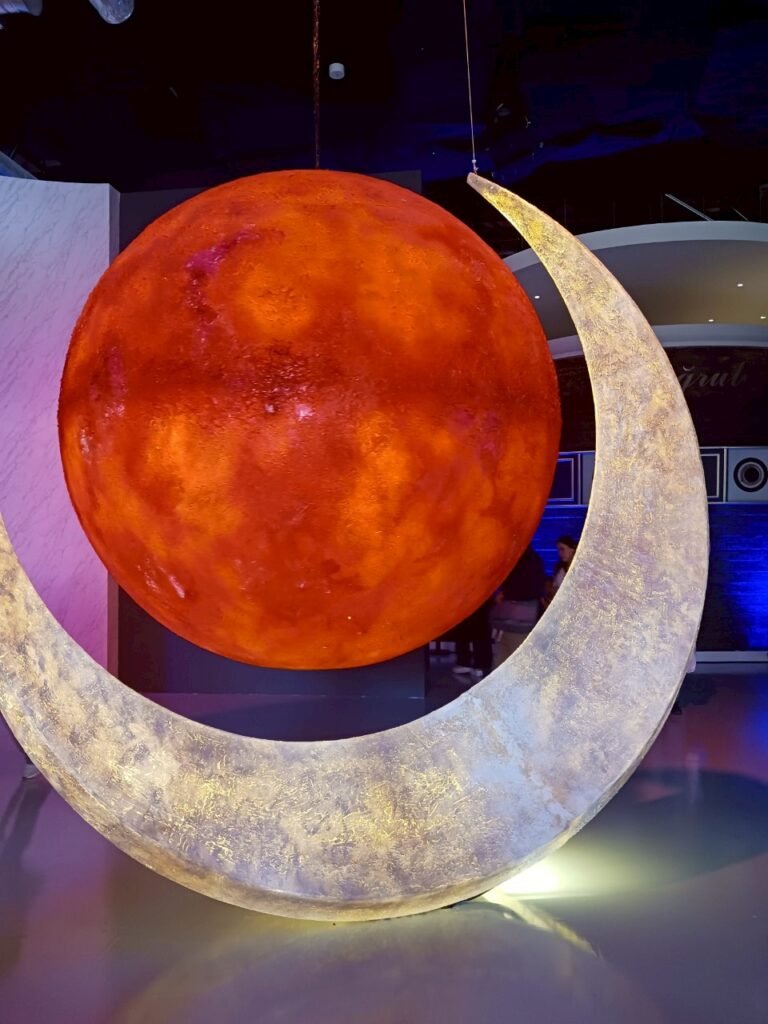 A suspended crescent moon and fiery planet sculpture in a dimly lit exhibit.