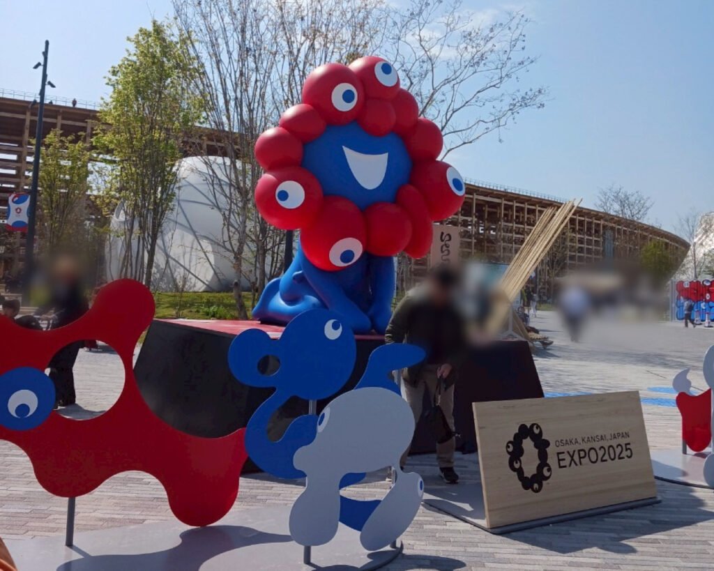 Statue of the Expo 2025 Osaka mascot with playful figures and a wooden sign
